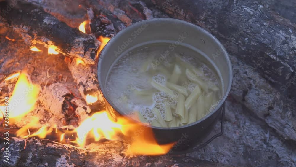 Cooking pasta with cauldron on campfire in evening forest. The cook stirs the food with a spoon. Close-up Cooking pasta with cauldron on campfire in evening forest. The cook stirs the food with a spoon. Close-up