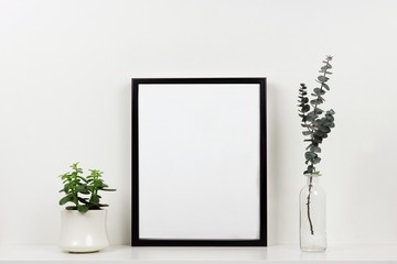 Mock up black frame with plant and branches on a shelf or desk. White shelf and wall. Portrait frame orientation.