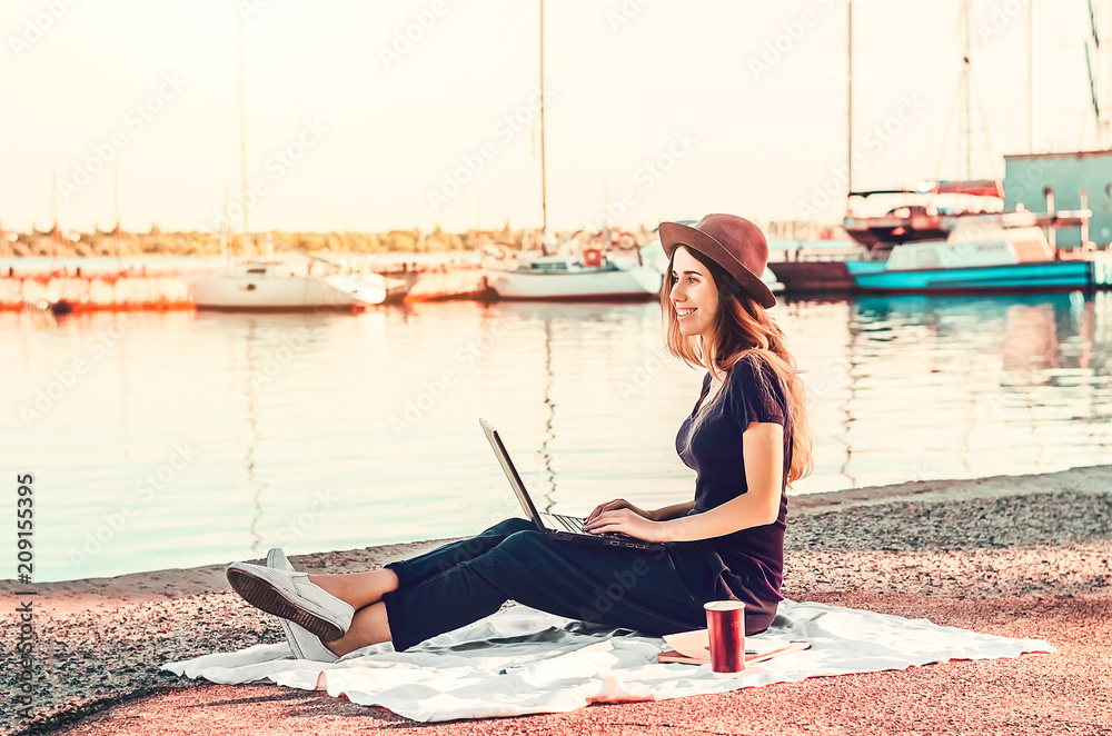 young girl sitting on the beach. hat on the head. the girl behind the laptop works wirelessly. hot coffee.