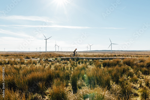 A woman riding a mountain bike in over a wooden track surrounded by wild grass at a wind farm in Scotland photo