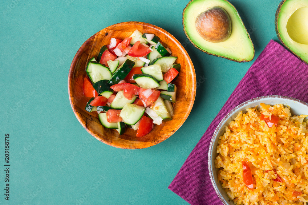 Traditional National Mexican Tomato Rice Stewed Pilaf with Hot Chili Peppers Garlic in Turquoise Bowl. Fresh Cucumber Onion Salsa Salad Avocado for Guacamole. Green Background. Top View. Copy Space