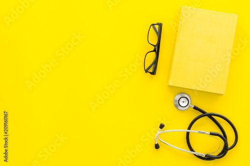 Medical literature. Stethoscope near book and glasses on yellow background top view copy space