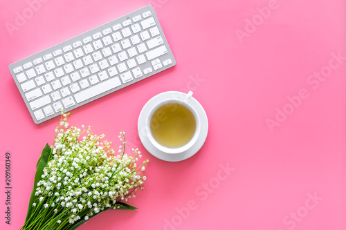 Cosy spring work desk concept. Computer keyboard  cup of tea and bouquet of lily of the valley flowers on pastel pink background top view copy space