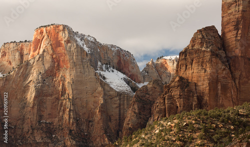 Horizontal view of The Alter of Sacrifice in Zion National park with it's red and yellow sandstone cliffs topped by snow and backed by a cloud filled sky with the sun breaking through