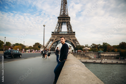 Photography Handsome man in dark pullover is standing on the bridge background of the Eiffel