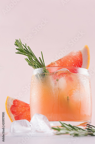 Refreshing cold citrus cocktail with ice cubes, green rosemary and slice grapefruit closeup on soft light pink and white background.