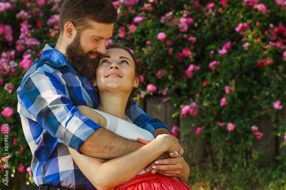 Romantic happy couple in love hugs and look at each other. attractive brunette woman and her handsome bearded boyfriend in park. Bush of roses flowers in the background Romantic happy couple in love hugs and look at each other. attractive brunette woman and her handsome bearded boyfriend in park. Bush of roses flowers in the background