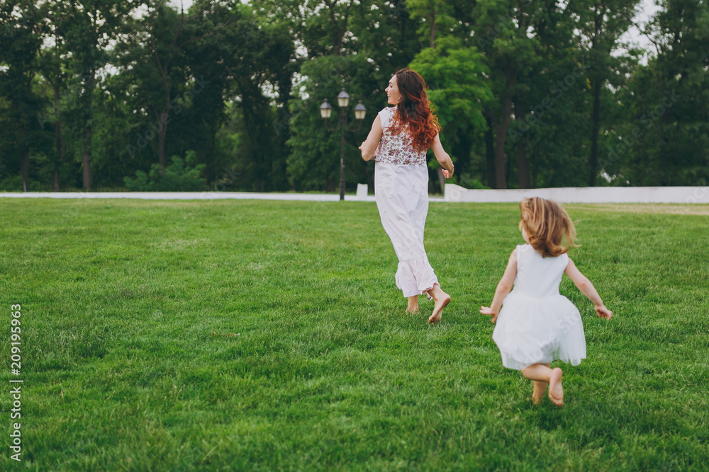 Little cute child baby girl running catching up have fun with joyful woman in light dress on green grass in park. Mother, little kid daughter. Mother's Day, love family, parenthood, childhood concept. Little cute child baby girl running catching up have fun with joyful woman in light dress on green grass in park. Mother, little kid daughter. Mother's Day, love family, parenthood, childhood concept.