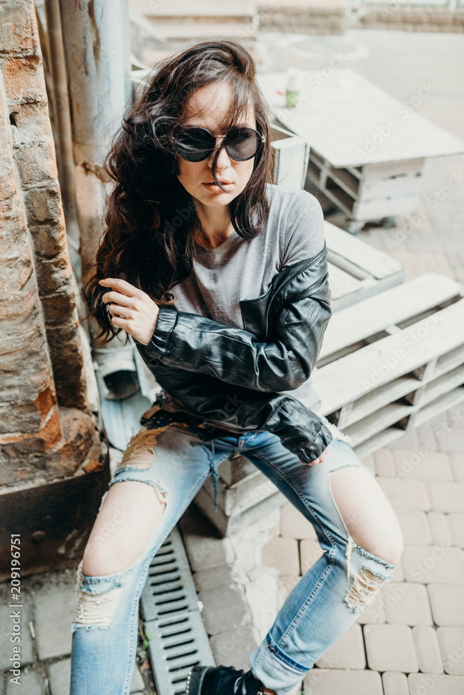 Confident and beautiful. Close up fashion woman, vintage denim biker  leather jacket, rock n roll style, fit girl posing on the street, stylish  outfit. On the background of light yard. Stock Photo