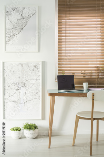 Maps on the wall next to a desk with a laptop and chair standing by the window with blinds in a workspace interior