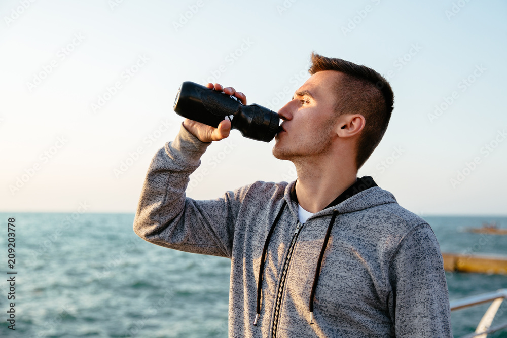 Obraz premium Side view of handsome sportsman drinking a water from bottle after running, workout, on quay, near the sea. Sport concept.