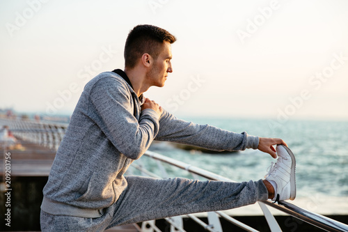 Young attractive man in sportswear on workout , doing exercises for legs, on quay, outdoors.