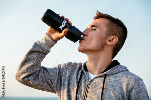 Side view of young sportsman drinking a water from bottle after running, workout, standing outdoors. Sport concept.