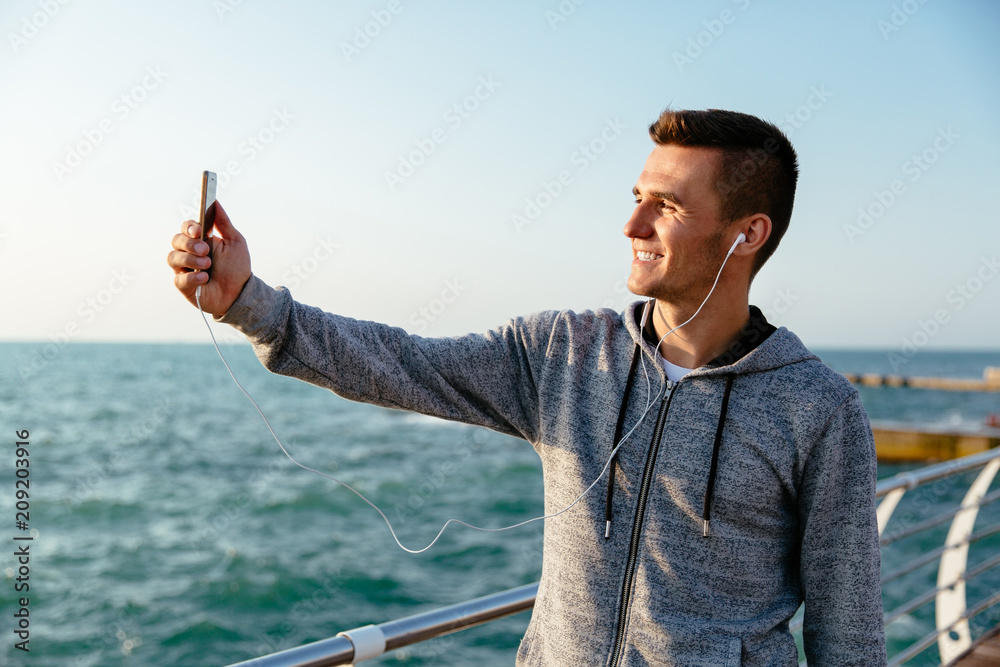 Fototapeta premium Smiling handsome man in headset and sport jacket takes a selfie on cellphone, while walking on pier. Outdoors.