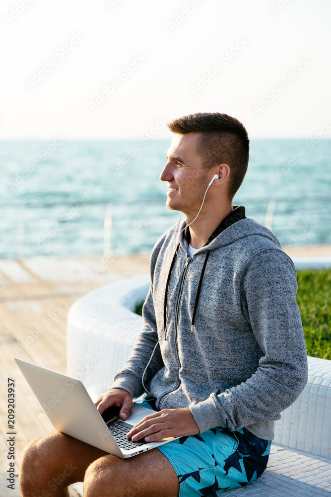 Smiling attractive man in headset, using a laptop, looking away, sitting on bench, on quay. Outdoors.