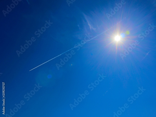 Bright sun with flares and spectral aureole around against blue sky with white jet traces