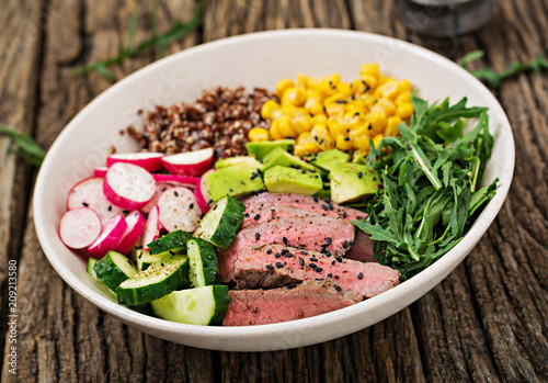 Healthy dinner. Buddha bowl lunch with grilled beef steak and quinoa, corn, avocado, cucumber and arugula on wooden background. Meat salad.