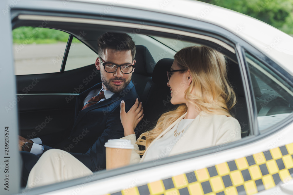 smiling young woman holding paper cup and flirting with handsome man sitting in taxi