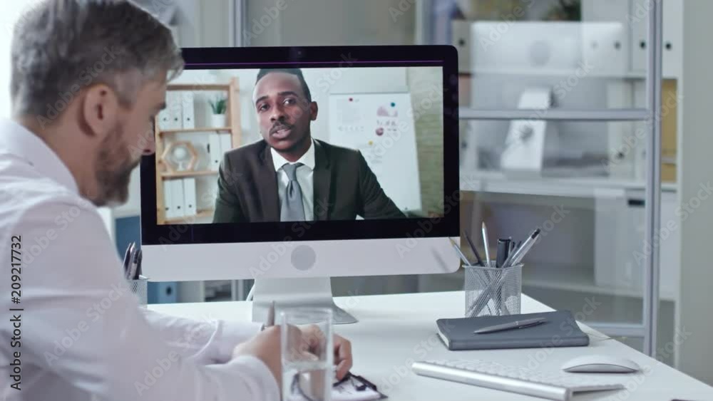 Mid-aged businessman sitting at office desk, writing down in notepad and discussing project with african american partner via video call on computer