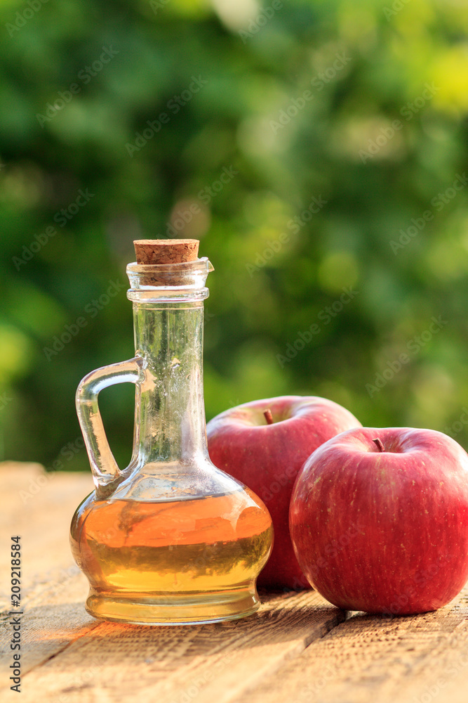 Apple vinegar in glass bottle with cork and fresh red apples on old wooden boards with blurred green natural background. Organic food for health Apple vinegar in glass bottle with cork and fresh red apples on old wooden boards with blurred green natural background. Organic food for health