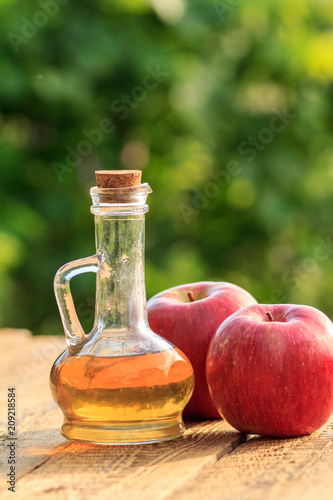 Apple vinegar in glass bottle with cork and fresh red apples on old wooden boards with blurred green natural background. Organic food for health