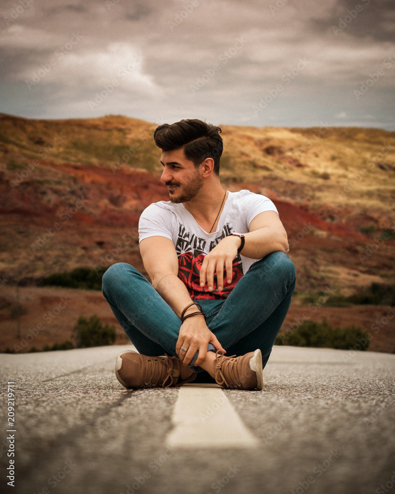 Smiling young man sitting on street in summer with legs crossed and looking to the side // Junger Mann auf Strasse im Valley / Wüste sitzend und lächelnd Smiling young man sitting on street in summer with legs crossed and looking to the side // Junger Mann auf Strasse im Valley / Wüste sitzend und lächelnd