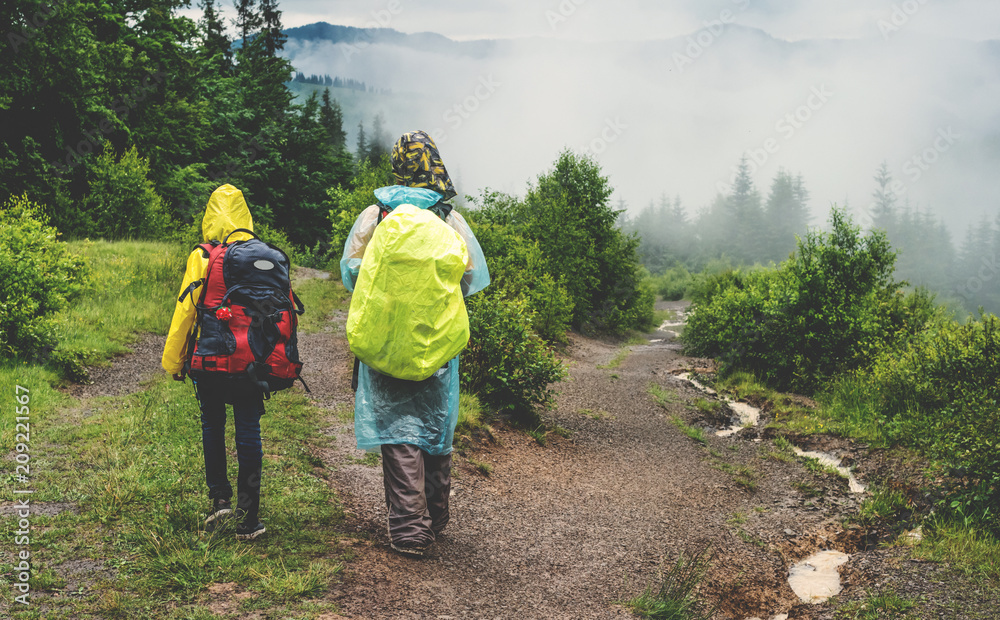 Two hikers tourist in raincoat walking on trail to green mountain forest in the fog with the yellow backpack in rainy weather. Two hikers tourist in raincoat walking on trail to green mountain forest in the fog with the yellow backpack in rainy weather.