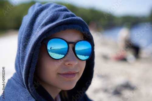 Closeup of pensive little girl in a hood and sunglasses looking away in thoughts with reflection of fisherman with fishing rod