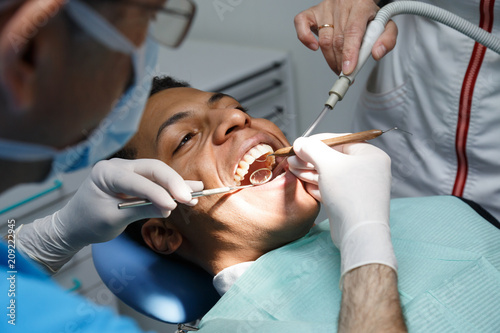 Dentist examining oral cavity of young African-American man working in in dental clinic with assistant. 