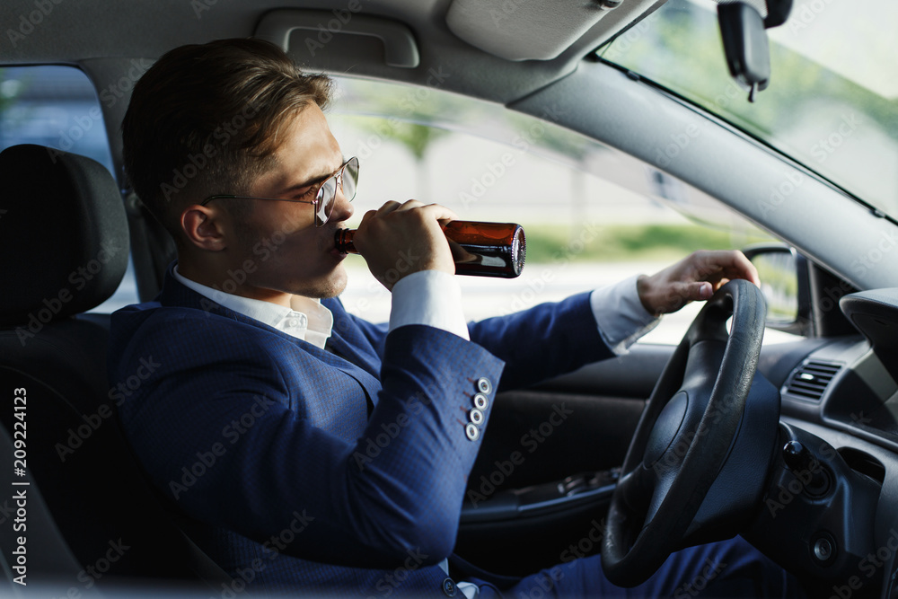 Handsome young businessman drinks beet sitting at the steering wheel inside the car