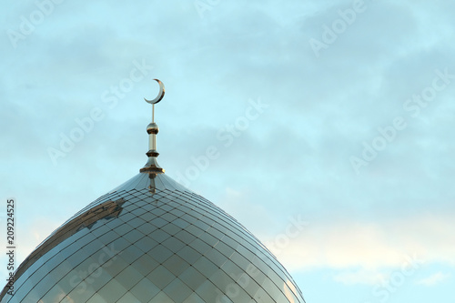 The symbol of Islam is a golden crescent moon on top of the mosque minaret on the blue morning sky with clouds.