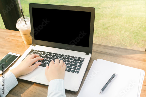 Close-up of business female working with laptop with blank black screen make a note document and smartphone in coffee shop like the background.