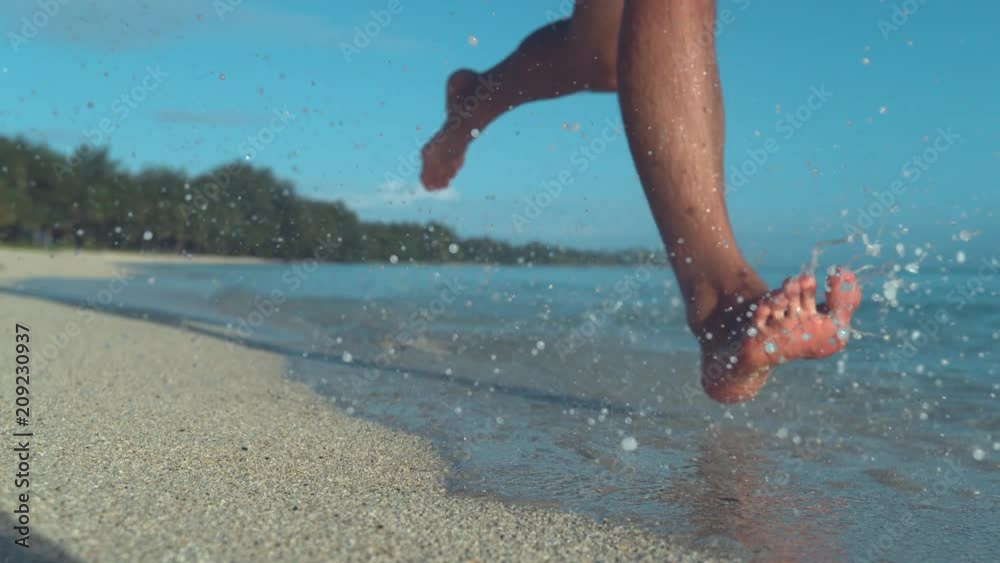 SLOW MOTION, LOW ANGLE: Glassy drops of sea water fly around unrecognizable man's feet as he runs down the sunny beach. Cool shot of male jogging along the white sand coastline of tropical island.