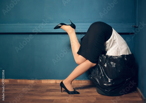Woman trying to get her head out of garbage basket. Lady dressed in business casual clothes got her head stuck in black trash can.