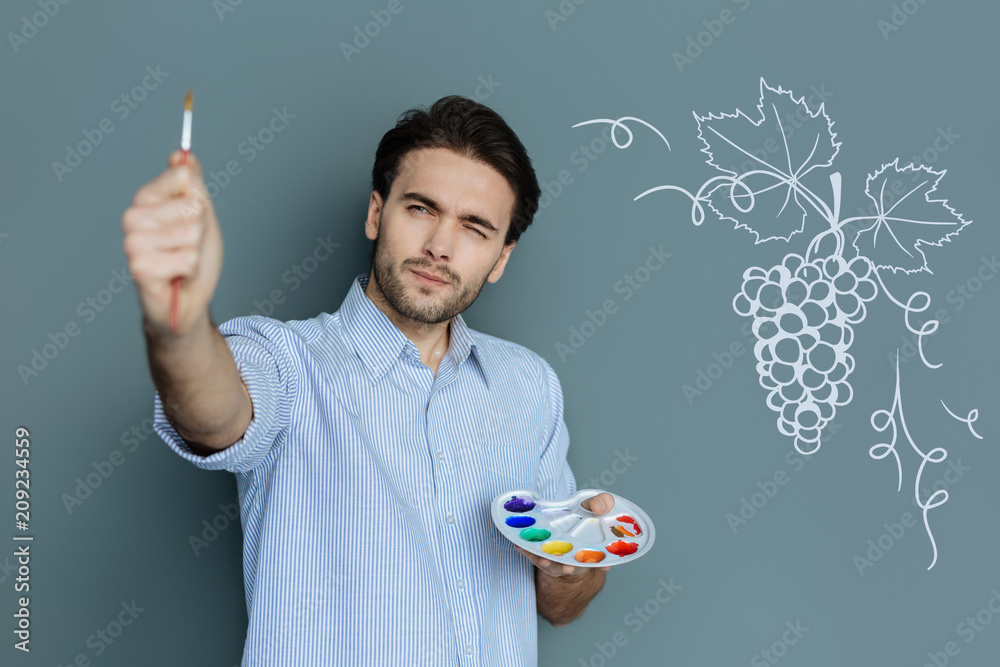 Attentive painter. Calm professional painter holding a color palette and thoughtfully looking at the brush in his right hand Attentive painter. Calm professional painter holding a color palette and thoughtfully looking at the brush in his right hand