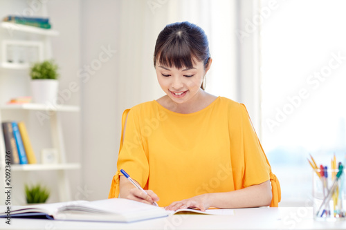 people  education  high school and learning concept - happy asian young woman student with book and notepad writing at home