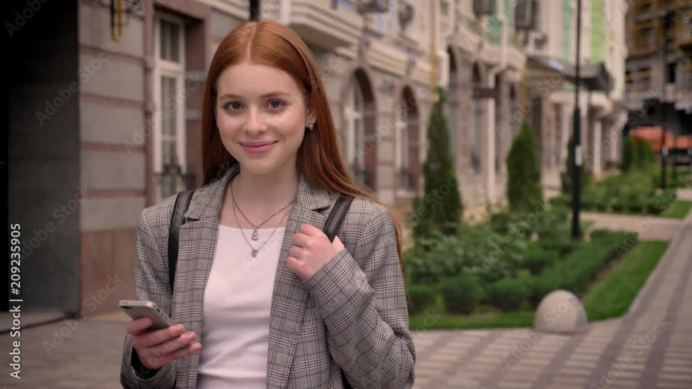 Young ginger charming woman standing in street, looking at phone and in camera, holding harness of backpack, smiling, building background Young ginger charming woman standing in street, looking at phone and in camera, holding harness of backpack, smiling, building background