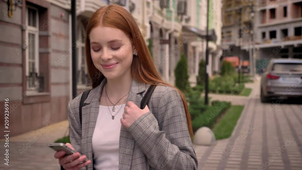 Young ginger woman standing in street, texting on phone and looking in camera, smiling, building background Young ginger woman standing in street, texting on phone and looking in camera, smiling, building background