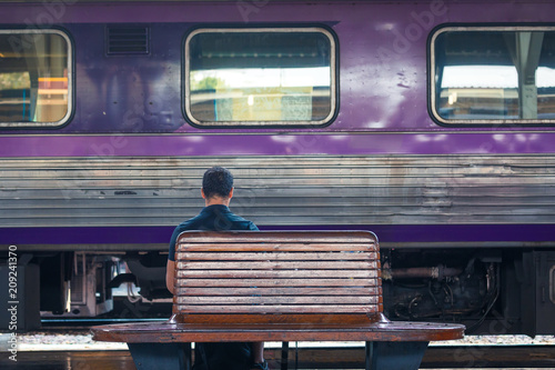 back of a man waiting on a bench at railway station with purple train in background during day