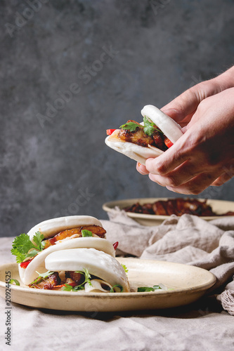 Man's hands hold asian sandwich steamed gua bao buns with pork belly, greens and vegetables served in ceramic plate on table with linen tablecloth. Asian style fast food dinner.