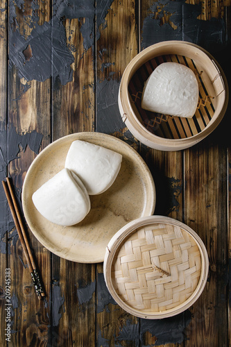 Empty gua bao steamed buns in ceramic plate and opened bamboo steamer over dark wooden plank background. Flat lay, space. Asian fast food.