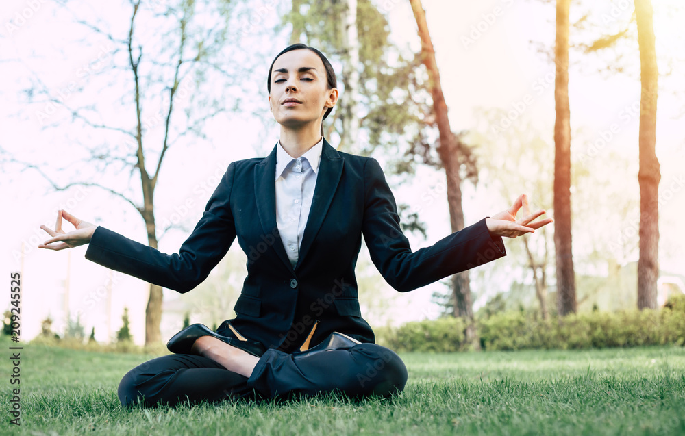 Young beautiful modern business woman in a suit meditating in a park sitting in a lotus position in the fresh air Young beautiful modern business woman in a suit meditating in a park sitting in a lotus position in the fresh air