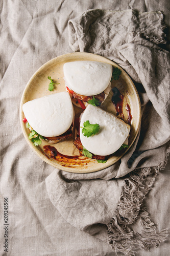 Asian sandwich steamed gua bao buns with pork belly, greens and vegetables served in ceramic plate over linen tablecloth. Asian style fast food dinner. Flat lay, space