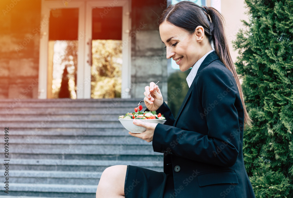 Healthy nutrition on break. Modern smiling business woman in a black suit with a fork eating a vegetable salad outdoors Healthy nutrition on break. Modern smiling business woman in a black suit with a fork eating a vegetable salad outdoors
