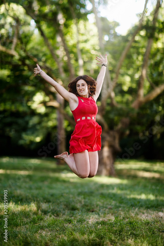 Happy girl jumping for joy and raising her arms. In the background the trees and the green woods of the park. Girl with freckles and red hair, wearing a short red dress.