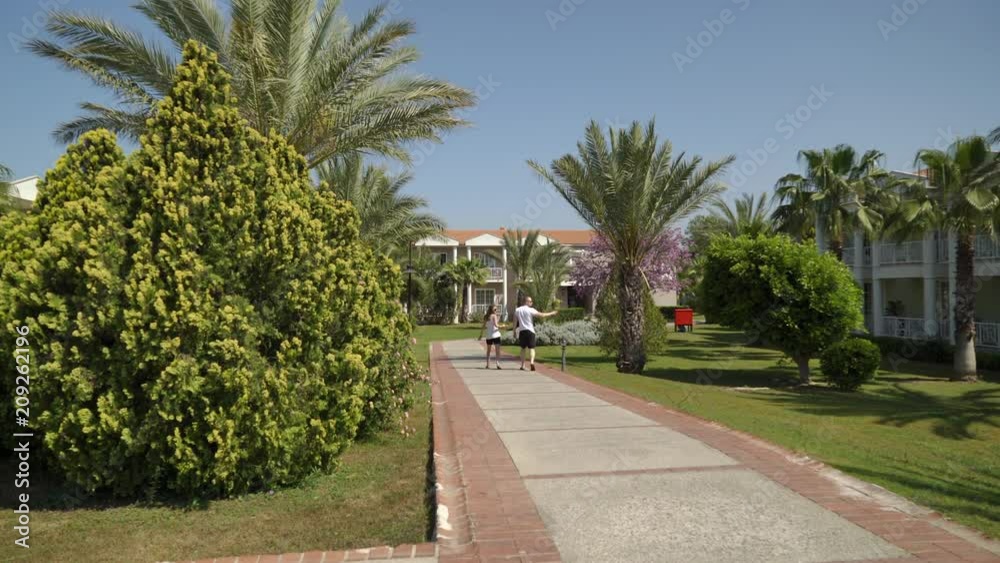 A young couple in white T-shirts walks along a path between palms and small buildings. A young couple in white T-shirts walks along a path between palms and small buildings.
