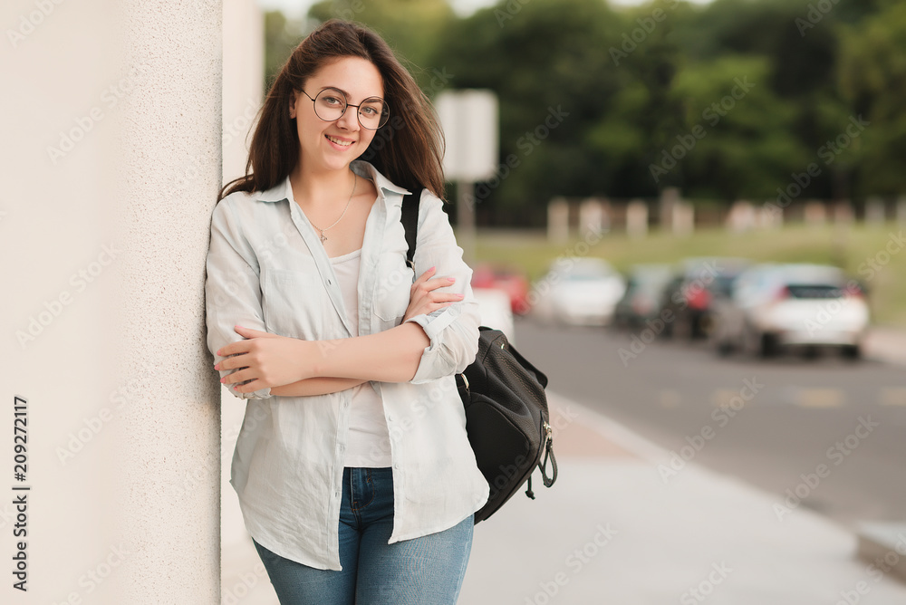 Portrait of a student girl with glasses. She stands in the background of the park and holds notebooks and folders in her hands. The girl is preparing to enter the university