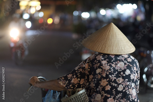 An elderly woman in a traditional Vietnamese straw hat walks with a bicycle through the night city