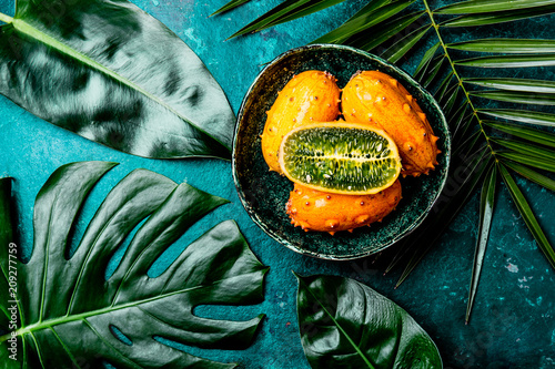 Fototapeta Naklejka Na Ścianę i Meble -  Tropical fruin KIWANO passion fruit in green bowl on turquoise background with tropical palm tree leaves. Top view. Tropical concept