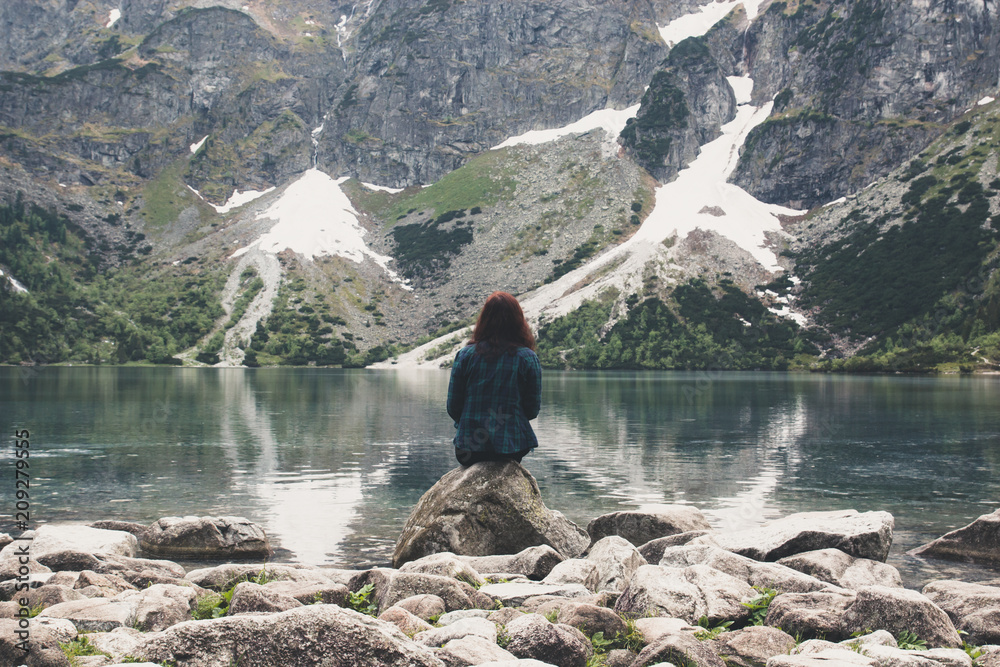 Young lonely brunette girl sitting in a romantic sadness on the shore of a blue lake in the mountains